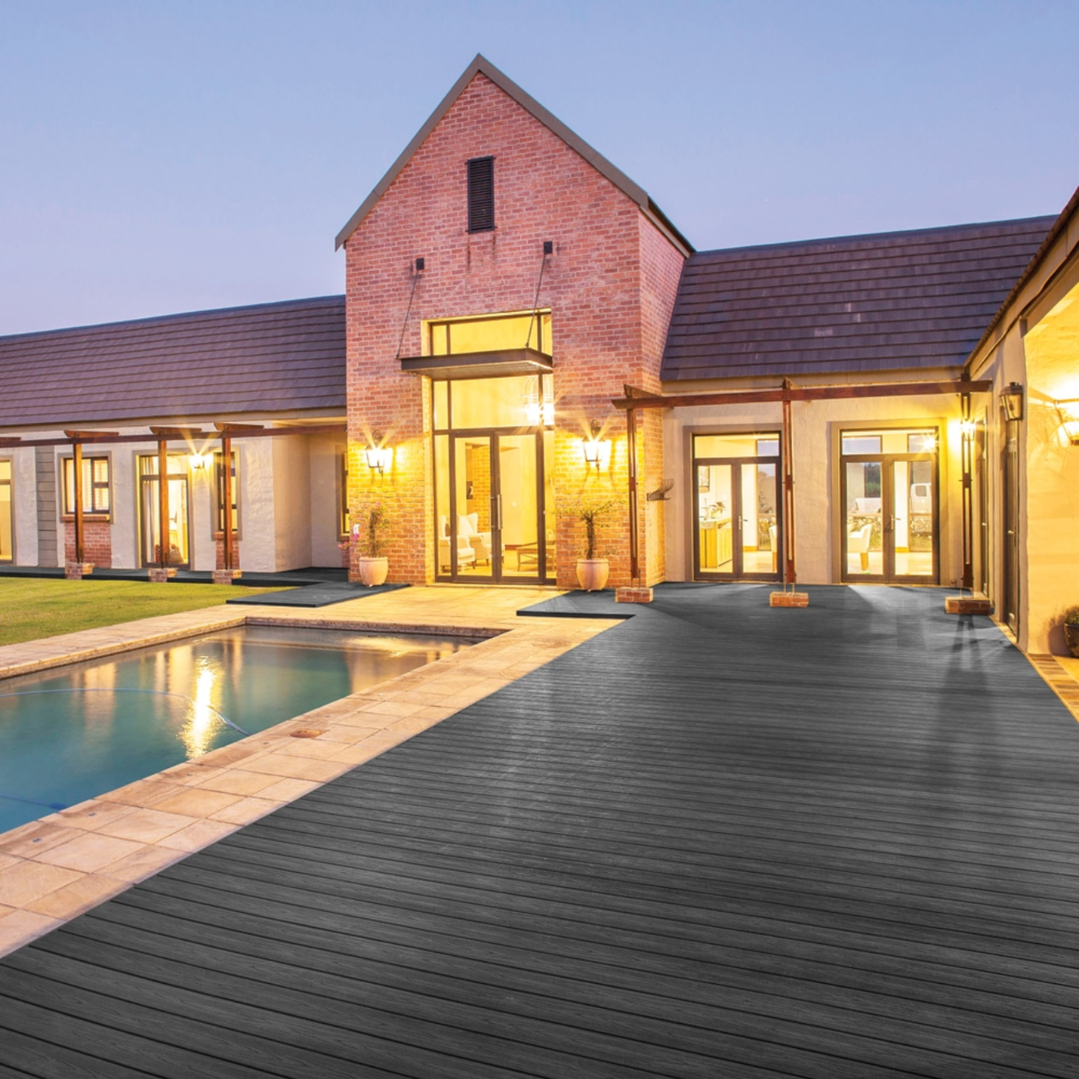 A modern brick house at dusk, featuring a large Eva-Last Infinity Baltic Nero composite deck next to a swimming pool, illuminated by warm outdoor lighting.