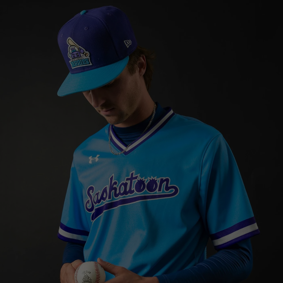 Person wearing a blue 'Saskatoon' baseball jersey and cap on a dark background