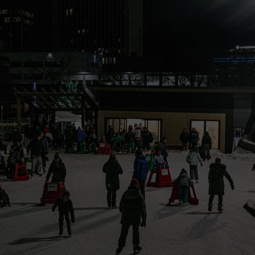 People skating on an outdoor ice rink at night with buildings in the background.